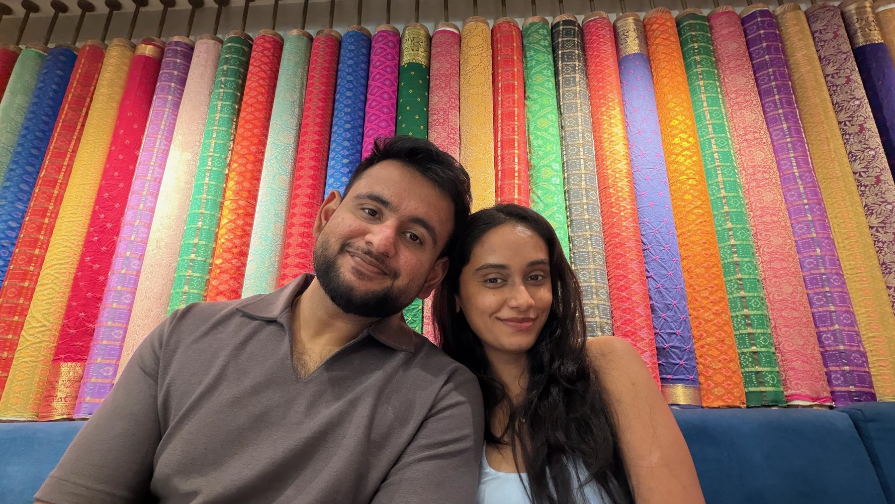 Yashika and Amol smiling together in front of colourful Indian textiles